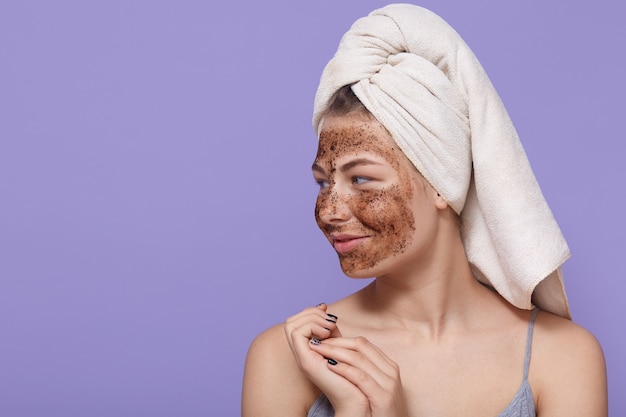 Portrait of female model applies chocolate mask on face, has positive expression, looks aside