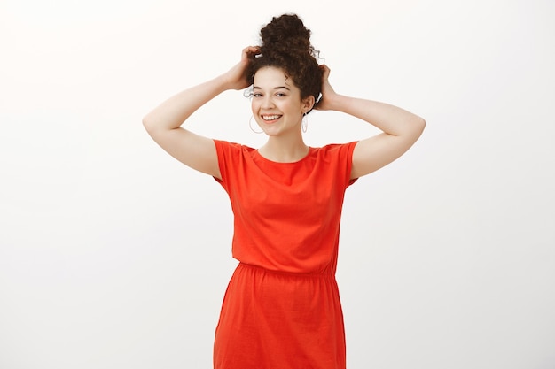 Portrait of femenine confident cute woman in stylish dress, touching combed hair and smiling with tender shy expression