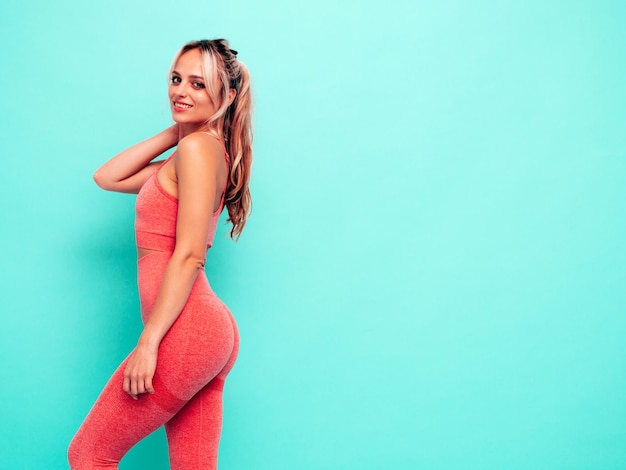 Portrait of fitness smiling woman in pink sports clothing Young beautiful model with perfect bodyFemale posing near blue wall in studioCheerful and happy Stretching out before training