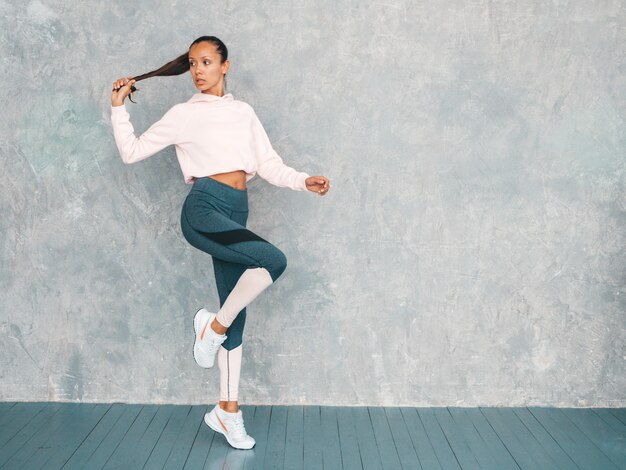 Portrait of fitness woman in sports clothing looking confident.Young female wearing sportswear.Beautiful model with perfect tanned body.Female jumping in studio near gray wall.Holds in hand hair tail