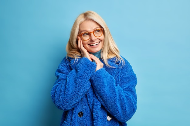 Free photo portrait of forty years old woman with blonde hair brilliant toothy smile keeps hands near face looks happily, has dreamy expression