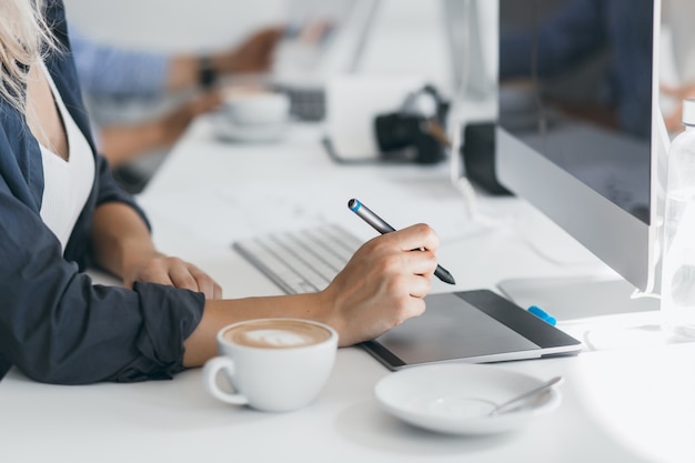 Portrait of freelance web-designer drinking coffee at workplace and holding stylus. Lightly-tanned lady in black shirt using tablet in her office, sitting in front of computer.