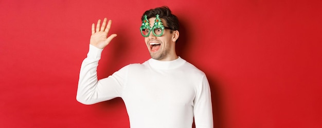 Free photo portrait of friendly cheerful man in party glasses and white sweater saying hello and looking left g...