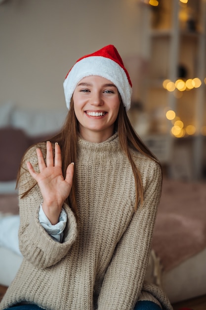 Portrait of friendly kind brunette woman in santa hat waving raised hand and saying hi to camera, enjoying christmas time.