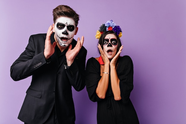 Free photo portrait of frightened and shocked boy and girl. couple with face art on day of all dead in dark attires posing on lilac wall.