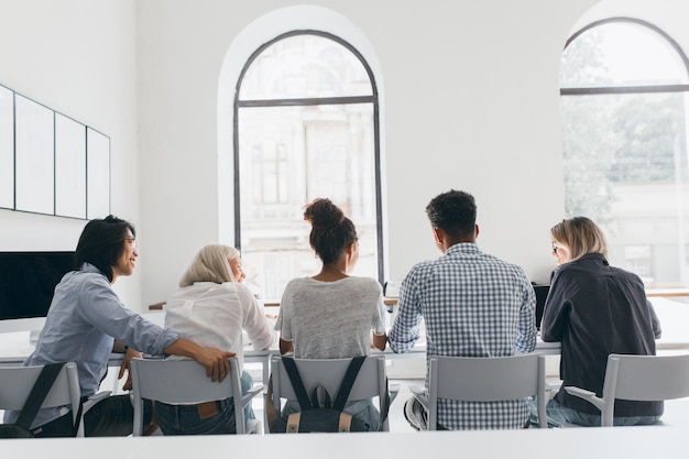 Portrait from back of african girl in white shirt with backpack resting with friends after lessons. Indoor photo of young people discussing something in conference hall with big windows.