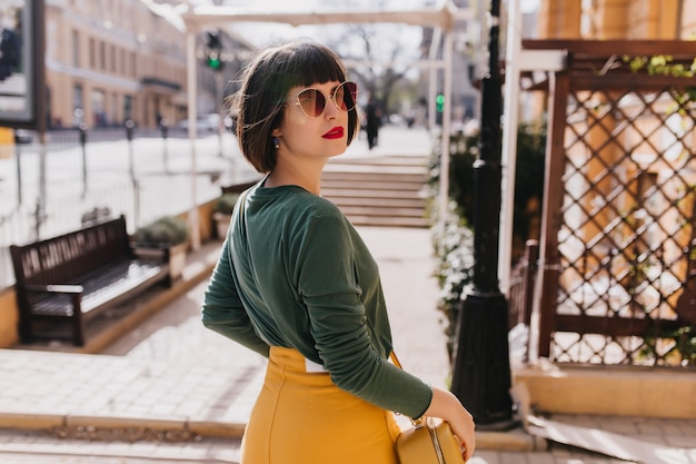 Free photo portrait from back of sensual brunette woman with red lips looking over shoulder. outdoor portrait of stylish european lady in green sweater walking down the street.