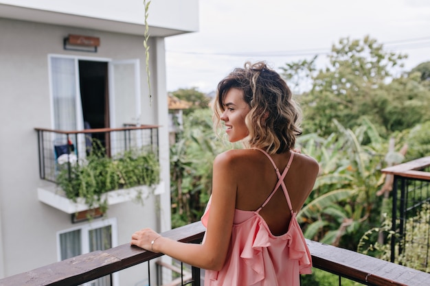 Portrait from back of stunning tanned woman in pink attire looking at trees. Outdoor photo of romantic blonde girl standing at balcony and enjoying city view.