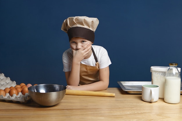 Portrait of frustrated 10 year old boy in chef uniform covering mouth feeling puzzled while going to make pancakes by himself for the first time with milk