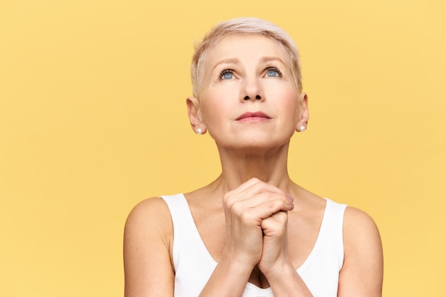 Portrait of frustrated nervous middle aged woman with short blonde hair looking up and holding hands clasped, having hopeful facial expression, praying to God, asking to help her through hard times