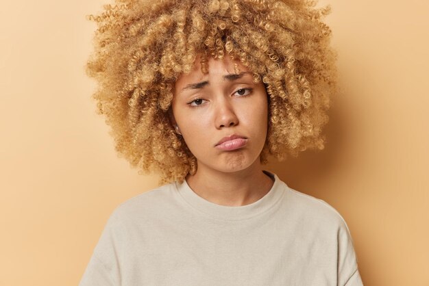 Portrait of frustrated sad curly woman looks unhappily feels sorry about what happened purses lips dressed in casual t shirt isolated over brown background People and negative emotions concept