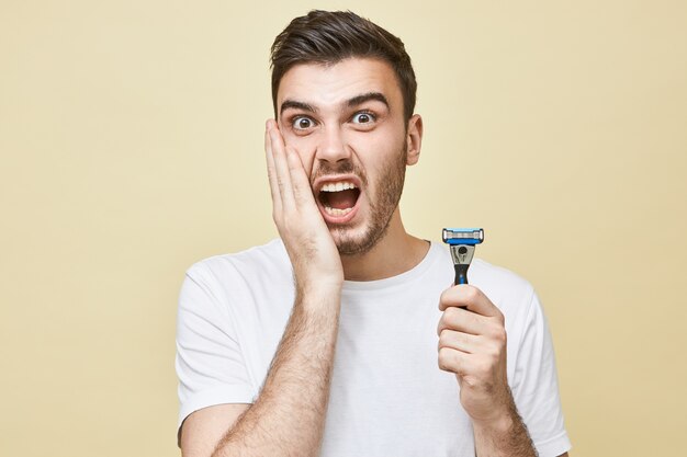 Portrait of frustrated unhappy young European man with stubble holding hand on his cheek and screaming, having terriified look, suffering from skin irritation because of shaving, holding razor stick