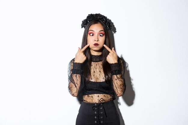 Portrait of funny asian girl animator on halloween holiday making faces, grimacing and poking cheeks, standing over white background.