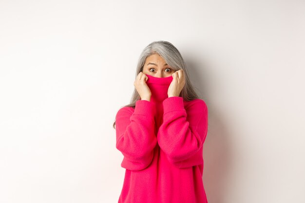 Portrait of funny asian grandmother hiding face in sweater collar, peeking at camera silly, standing over white background