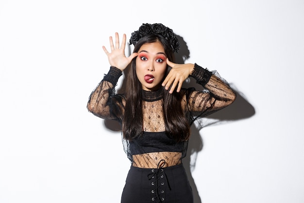 Free photo portrait of funny asian woman in witch costume celebrating halloween, mocking someone, showing faces and sticking tongue, standing over white background.