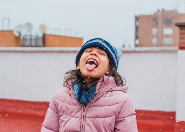 Portrait of a funny cute girl in a warm winter coat and hat catching snowflakes on her tongue