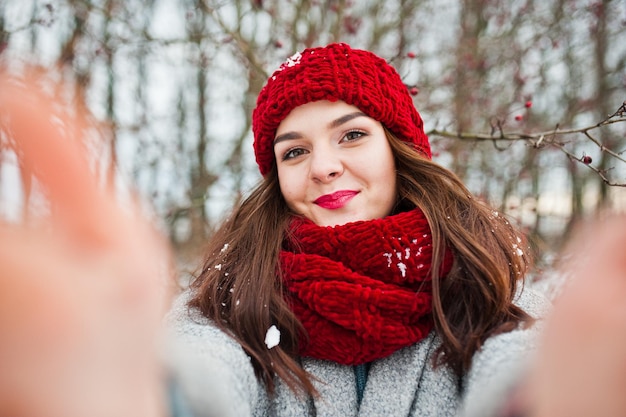 Free photo portrait of gentle girl in gray coat red hat and scarf near the branches of a snowcovered tree holding camera for selfie