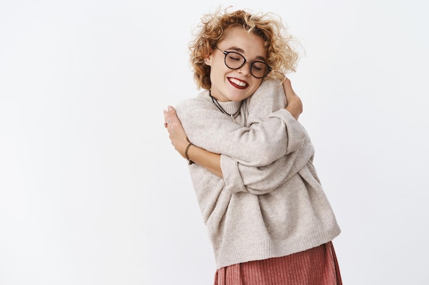 Free photo portrait of gentle and lovely beautiful woman in sweater and glasses leaning on shoudler huggng herself with hands over body and smiling sensually, feeling comfort.