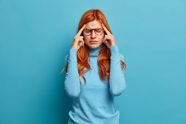 Free Photo portrait of ginger young european girl suffers severe headache keeps index fingers on temples tries to concentrate and continuing working dressed in casual clothes.