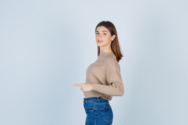 Portrait of girl pointing somewhere by standing sideways in sweater, jeans and looking cheery