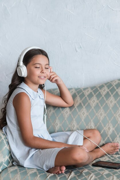 Portrait of a girl sitting on sofa enjoying the music on headphone attached to cell phone