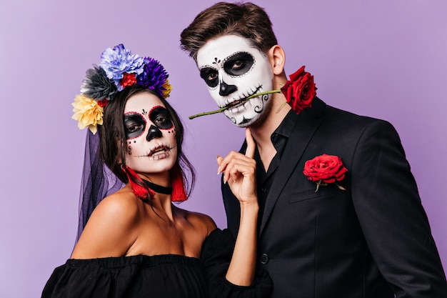 Free photo portrait of girl with flowers in dark hair touching painted face of passionate man with rose in his teeth.