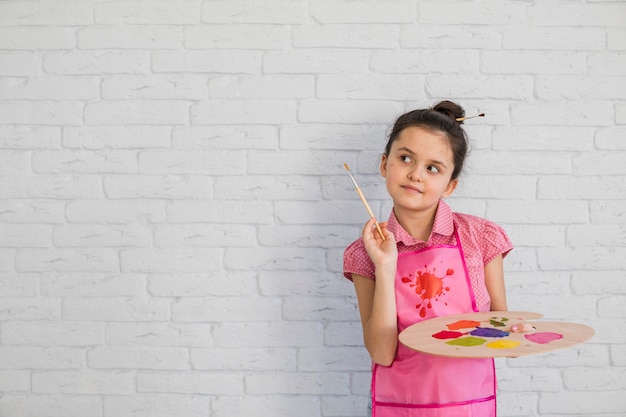 Free Photo portrait of a girl with paintbrush and palette standing against white wall