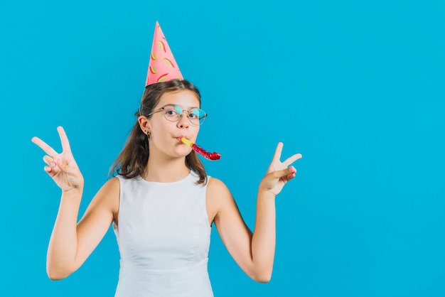 Free photo portrait of a girl with party horn gesturing peace sign on blue backdrop