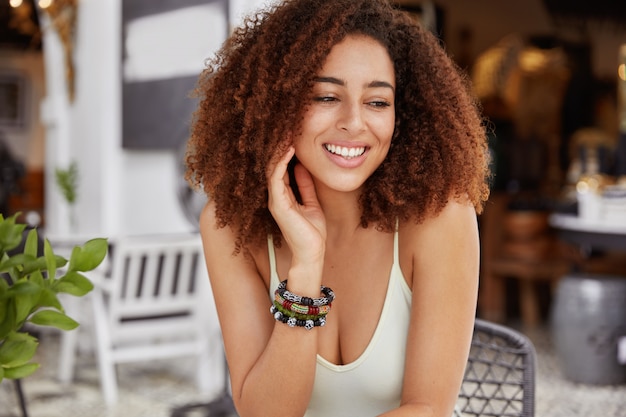 Portrait of glad African American female has positive smile, has bushy hairstyle looks with dreamy expression away sits against cafeteria interior.