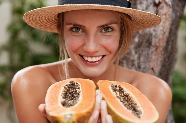 Free Photo portrait of glad lovely woman poses naked, wears summer hat, holds organic exotic papaya