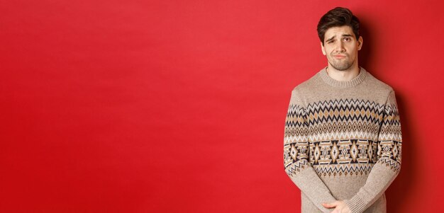 Portrait of gloomy and sad caucasian man in christmas sweater, frowning and pouting indecisive, having bad new year, standing over red background.