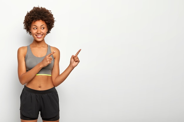 Portrait of good looking Afro woman points away on blank space, smiles pleasantly, wears top and shorts, copy space against white wall