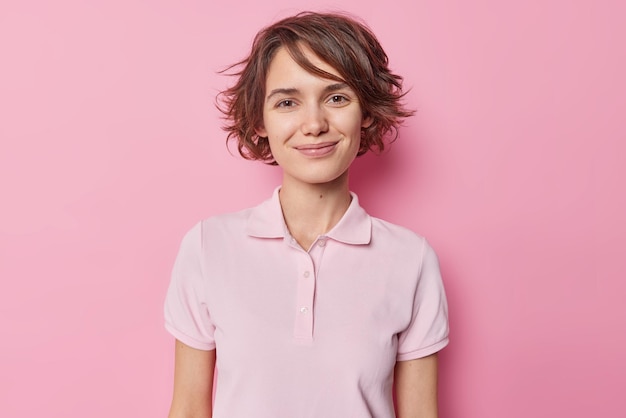 Portrait of good looking brunette woman smiles tenderly has charming look wears casual t shirt looks satisfied at camera isolated over pink studioo background receives pleasant news from interlocutor