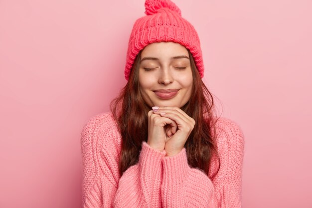 Portrait of good looking charming woman keeps both hands under chin, eyes closed, being deep in thoughts, models over rosy studio wall, dressed in sweater and hat