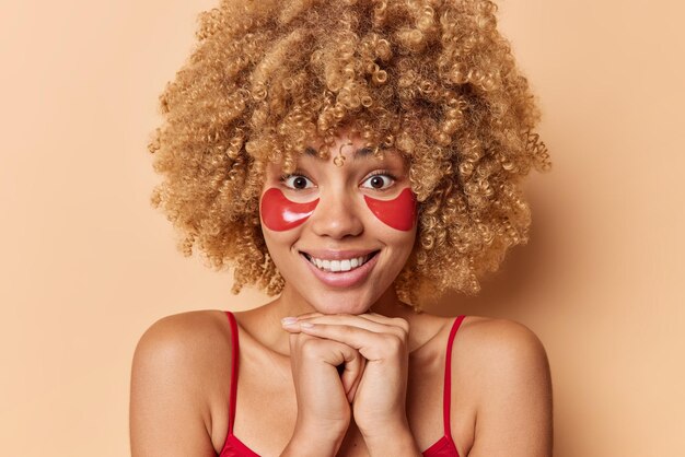 Portrait of good looking European woman with curly bushy hair smiles pleasantly keeps hands under chin applies red hydrogel patches under eyes dressed casually isolated over brown background