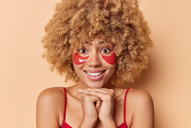 Portrait of good looking European woman with curly bushy hair smiles pleasantly keeps hands under chin applies red hydrogel patches under eyes dressed casually isolated over brown background