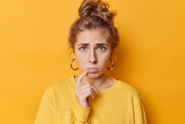 Free photo portrait of good looking frustraed young woman looks sadly purses lips has combed hair wears earrings and casual jumper isolated over yellow background negative emotions and feelings concept