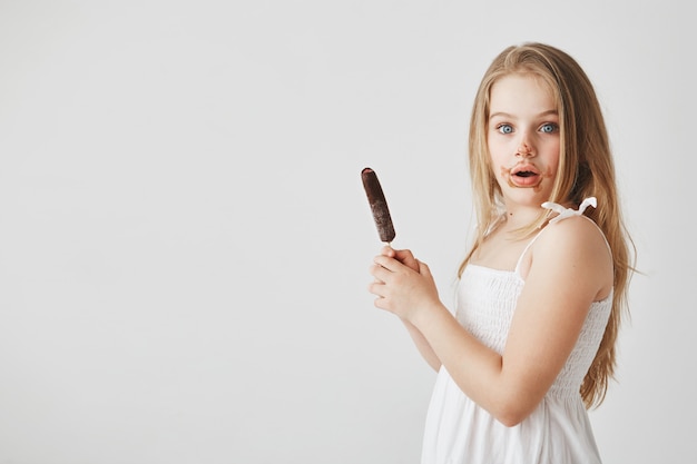 Portrait of good-looking funny small girl with blond long hair  with surprised face expression, being dirty after eating ice cream.