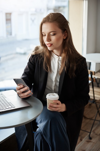 portrait of good-looking modern woman in cafe, sitting near window, drinking coffee and browsing in smartphone, having break of work with laptop