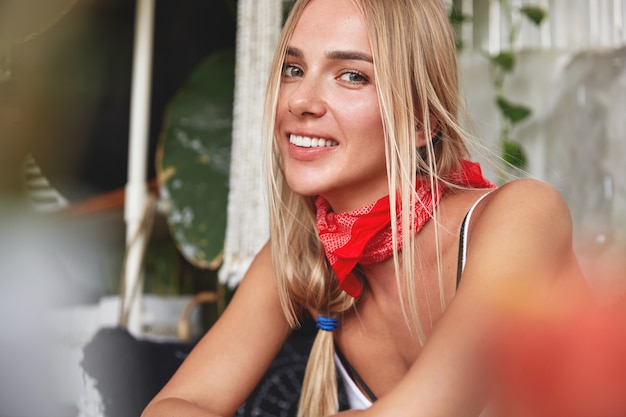 Portrait of good looking relaxed young female model with red bandana on neck, has her own style, sits against cafe cozy interior