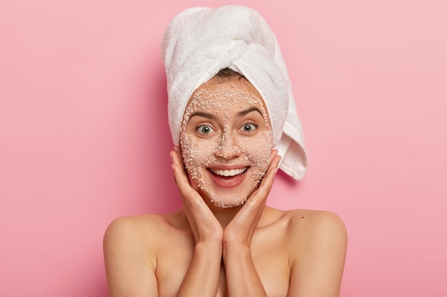 Portrait of good looking woman touches gently cheeks, has bare shoulders, smooth healthy skin, smiles pleasantly, applies peelig scrub on face, isolated over pink background, wears bath towel