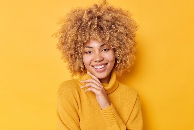 Free photo portrait of good looking young woman touches chin gently smiles gladfully looks with tender romantic expression wears warm jumper isolated over yellow background. pleasant human face expressions