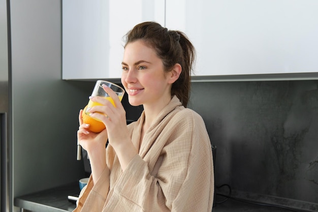 Free photo portrait of goodlooking woman in bathrobe drinks orange juice from glass and smiles starts her day