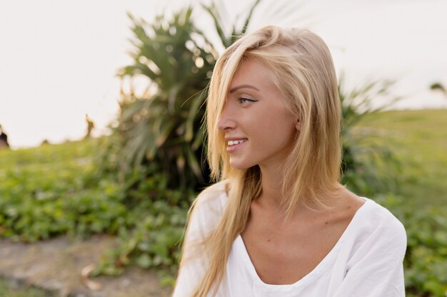 Portrait of gorgeous 20s european woman with long hair in summer white dress laughing and looking away while walking on the sand by the sea