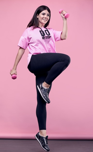 Portrait of a gorgeous body positive Latin woman in a pink sports hoodie exercising with dumbbells on pink