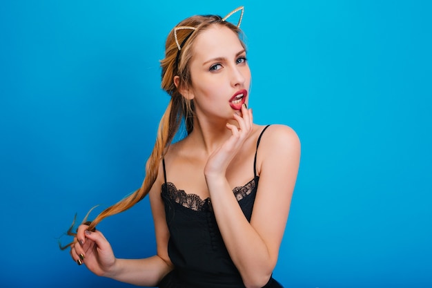 Free photo portrait of gorgeous girl with sexy look, touching her red lips at party. she has long blonde hair, nice manicure. wearing black dress, diadem with cat ears.