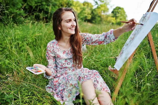 Free photo portrait of a gorgeous happy young woman in beautiful dress sitting on the grass and painting on paper with watercolors