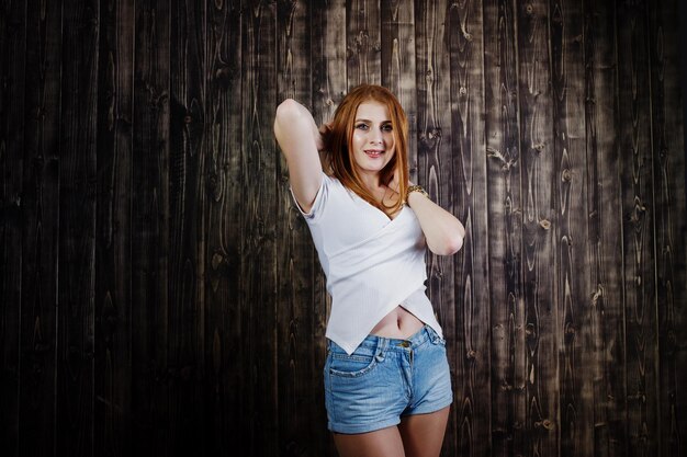 Portrait of a gorgeous redheaded girl in white tshirt and denim shorts posing in the studio next to the wooden wall