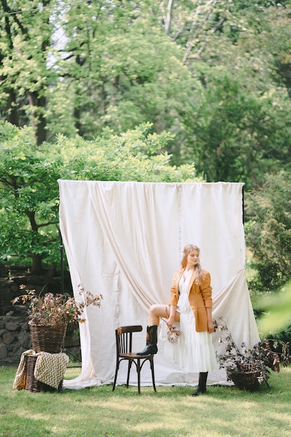 Free photo portrait of gorgeous woman at garden with white wall standing and looking in white dress and brown jacket, during daytime.