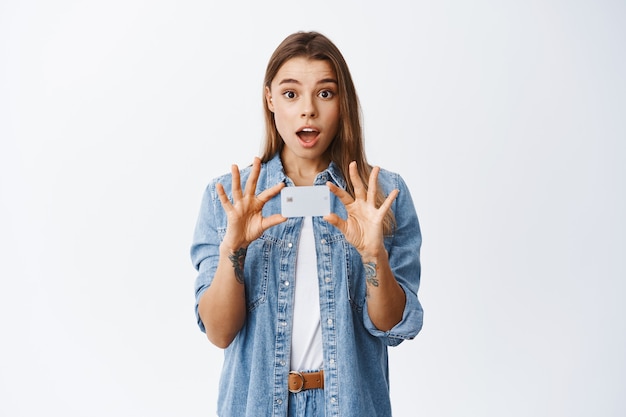 Portrait of gorgeous yougn woman in casual clothes introduce bank advertisement, showing plastic credit card and staring amazed at front, white wall